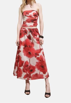 Vrouw die een rood-wit bloemig strapless top draagt en een bijpassende lange rok met zwarte open teen hakken en een zwarte armband aan haar rechterpols.