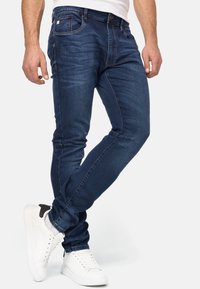 Vaqueros de mezclilla azul oscuro con un corte ajustado, que presentan sutiles variaciones en la textura y costuras en contraste. Se llevan con zapatillas blancas.