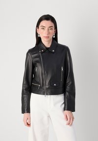 Een vrouw draagt een zwarte leren cropped jack met zilveren hardware, gecombineerd met witte broeken, terwijl ze tegen een lichtgrijze achtergrond staat.