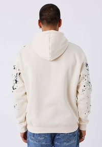 Man van achter gezien, gekleed in een crèmekleurige hoodie met zwarte verftroep-details op de mouwen en blauwe jeans met witte tekst.