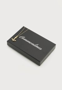 Κολιέ με χρυσό μενταγιόν άγκυρας που ξεκουράζεται πάνω σε μαύρο κουτί με την επιγραφή "Commonlines" σε λευκή καλλιγραφική γραμματοσειρά.