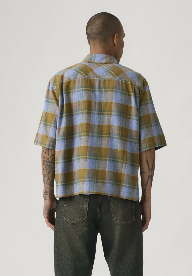 Man met een kort mouw geruit overhemd in blauw, geel en groene tinten, gecombineerd met donkergroene spijkerbroek, gezien van achteren.