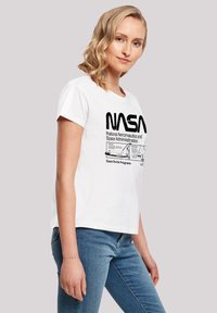 White short-sleeve t-shirt featuring black NASA logo and text. Casual fit with round neckline, made from soft cotton fabric. Blue jeans worn.