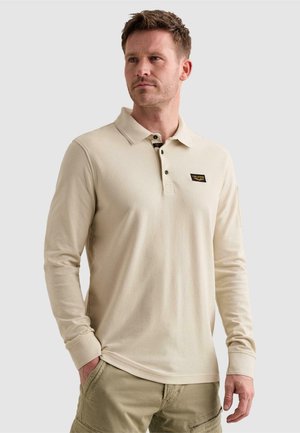 Homme portant un polo beige à manches longues avec des boutons noirs et un pantalon cargo kaki, debout avec une main dans la poche.