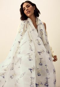 Balta šalle ar vīna glāžu un lentu rakstu, izgatavota no viegla, teksturēta auduma ar maigu krāsu.