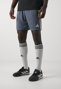 Grå idrettsshorts med svarte detaljer og hvite striper, kombinert med svarte fotballsko med tre hvite striper og knelange hvite sokker.