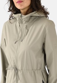 Chaqueta impermeable beige claro con capucha, cintura ajustada, cremallera frontal y cordones ajustables. Textura lisa y marca mínima.