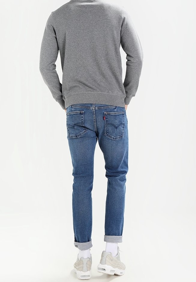 Sudadera gris, vaqueros azules con puños doblados, bolsillos traseros con detalles de costura y zapatillas claras, presentando un conjunto casual.