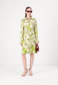Rochie crem cu un model floral verde strălucitor, mâneci lungi, talie marcată și lungime până la genunchi. Modelul poartă o mică geantă roșie și ochelari de soare.
