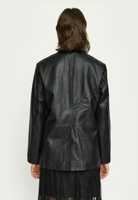 Zwart leren blazer met een gestructureerde silhouet, zachte textuur en naaddetails, gelaagd over een zwart kanten jurkje.