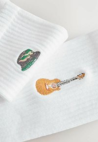 Bijele čarape s rebrastom teksturom, ukrašene zelenom kapom i smešnim gitarom u kontrastnim bojama.