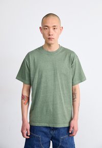 Grønn bomull T-skjorte med rund hals, korte ermer og avslappet passform; brukt med denimshorts; har synlige arm tatoveringer.