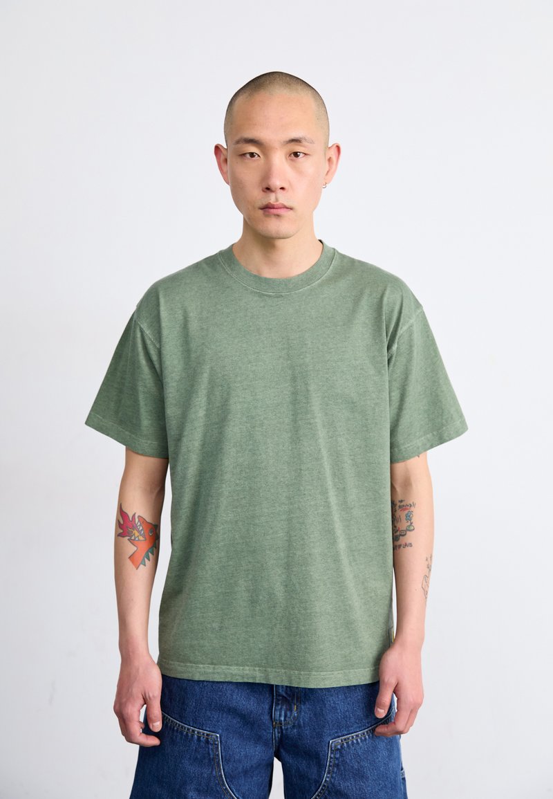 Grønn bomull T-skjorte med rund hals, korte ermer og avslappet passform; brukt med denimshorts; har synlige arm tatoveringer.