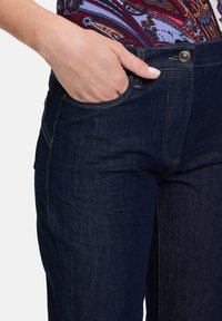 Jeans in denim blu scuro con una texture liscia, dotati di tasca frontale, chiusura a bottone metallico e dettagli di cuciture a contrasto.