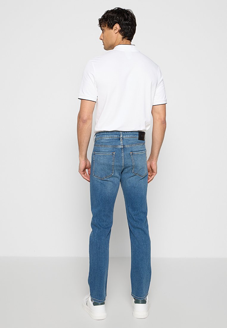 Homme portant un polo blanc, un jean bleu et des baskets blanches, se tenant de dos contre un fond uni.