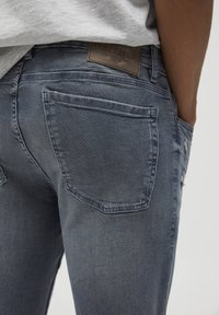 Vaqueros de mezclilla gris deslavado con una textura ligeramente desgastada, que presentan bolsillos traseros curvados y un parche de cuero marrón de la marca en la cinturilla.