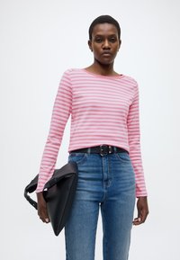 Μακρυμάνικη ροζ μπλούζα με οριζόντιες ρίγες, συνδυασμένη με ψηλόμεσα μπλε τζιν. Το μοντέλο κρατά μια μαύρη τσάντα με τριγωνικό σχήμα.