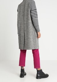 Grauer Fischgrät-langmantel mit Ärmeln aus schwarz-weißer Hahnentrittmuster, kombiniert mit rosa geraden Hosen und schwarzen Schnürstiefeln.