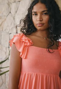 Robe rose corail avec un corsage smocké, des manches à volants dénudées des épaules et une jupe fluide, se détachant sur un fond de mur en pierre.