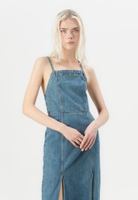 Farmernadrág anyagú, vékony pántos nyári ruha, négyzet alakú dekoltázzsal és oldalsó sliccekkel; megjelenése elhalványult kék mosású és egyenes szabású.