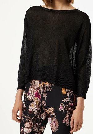 Pull noir transparent à manches longues porté avec un pantalon noir à fleurs roses, jaunes et beiges, montré sur une personne de l'épaule aux hanches.