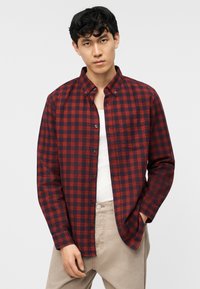 Νεαρός άνδρας που φοράει κόκκινο και μαύρο καρό πουκάμισο πάνω από ένα λευκό μπλουζάκι, με μπεζ παντελόνι, στέκεται μπροστά σε απλό φωτεινό φόντο.
