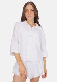Weiße, leichte Baumwollbluse mit Knopfverschluss, kurzen Ärmeln, strukturiertem Stoff und einer Brusttasche. Kombiniert mit passenden, rüschendetails Shorts.