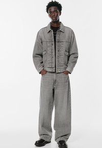 Mladic stoji s rukama u džepovima, odjeven u sivi traper jaknu, široke sive traperice, crnu majicu, slojevite srebrne lance i crne čizme.