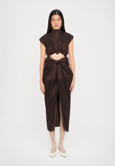 Braun plissiertes Kleid mit Knotendesign an Taille und Schultern, offenen Seiten und einem Schlitz vorne. Das Model steht vor einem neutralen Hintergrund.