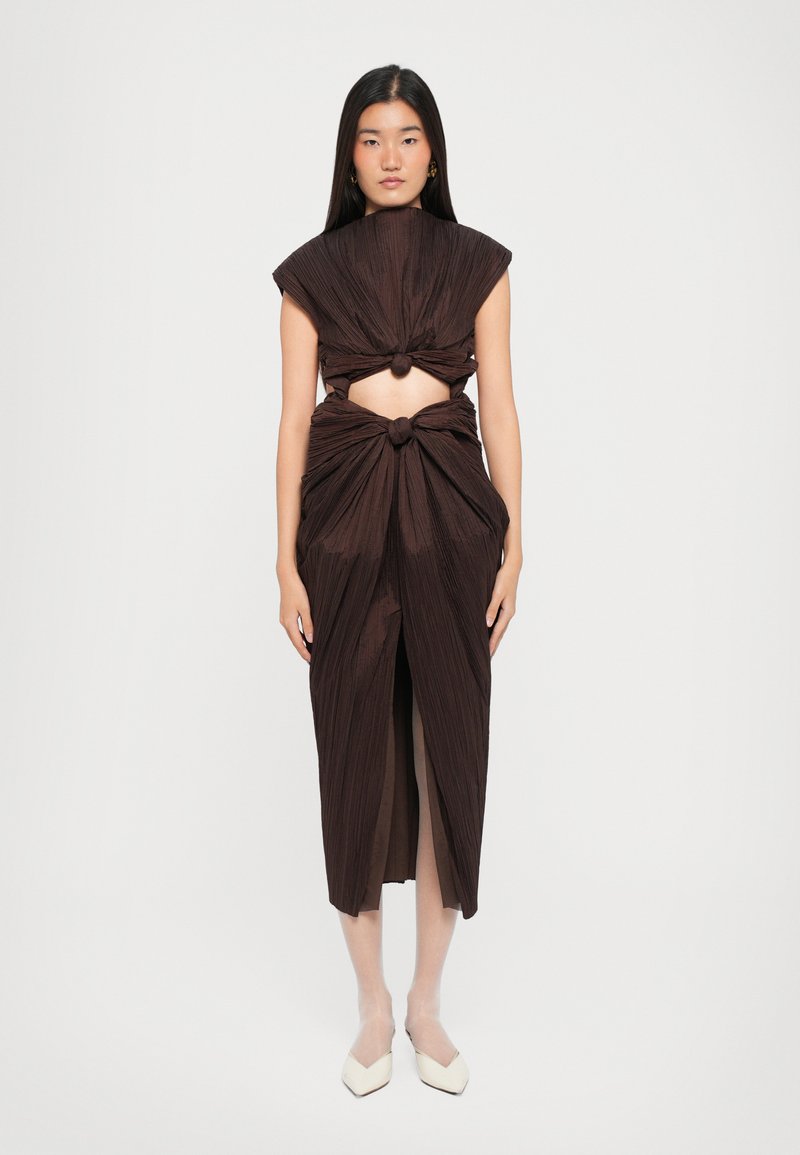 Robe plissée marron présentant un design en nœud à la taille et aux épaules, avec des côtés ouverts et une fente à l'avant. Le mannequin se tient devant un fond neutre.