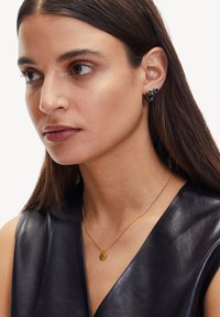 Zwarte spiraaloorbellen en een metalen ketting met een gouden hanger die door een model worden gedragen. De top van het model is een mouwloze, diepe V-hals leren ontwerp.