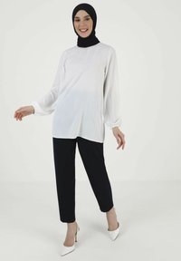 Weißes Langarmoberteil mit gerafften Manschetten, kombiniert mit schwarzen Hosen. Das Model trägt ein schwarzes Hijab und weiße High Heels. Einfaches, elegantes Design.