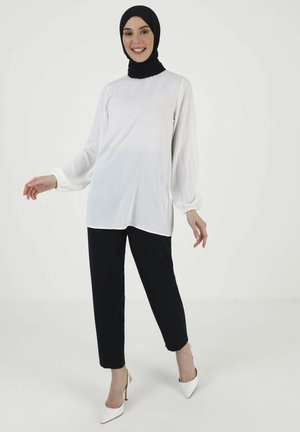 White long-sleeve top with gathered cuffs, paired with black trousers. Model wears a black hijab and white high-heeled shoes. Simple, elegant design.