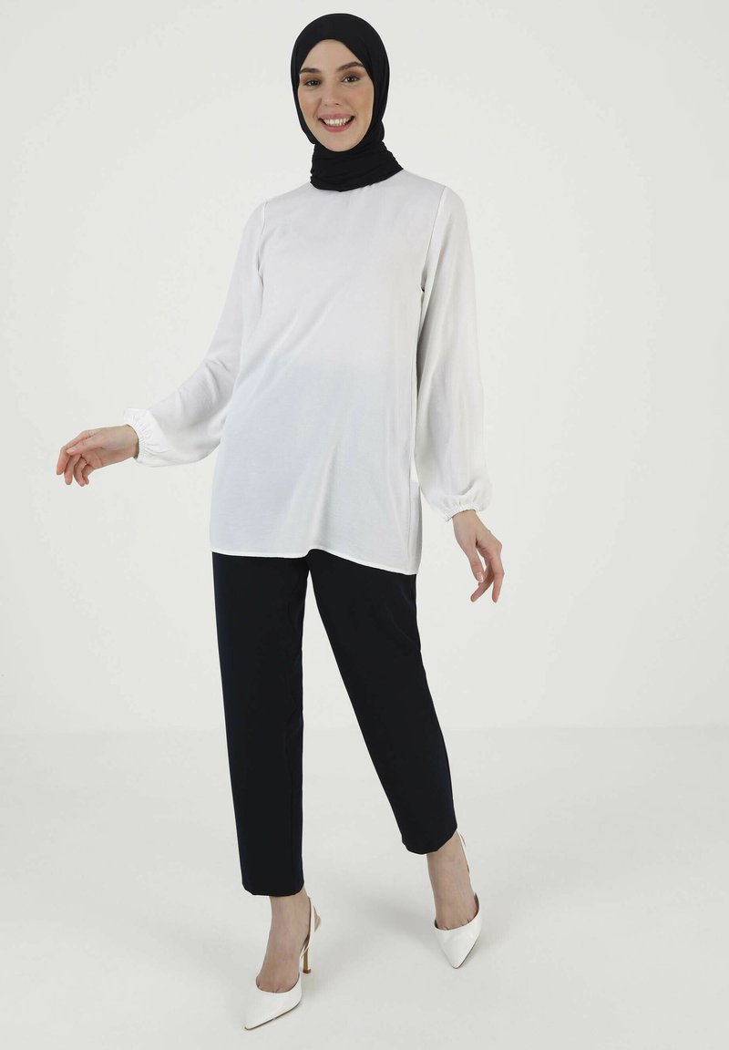 Vit långärmad topp med rynkade manschetter, ihopparad med svarta byxor. Modellen bär en svart hijab och vita högklackade skor. Enkelt, elegant design.