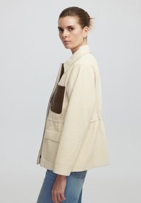 Crème gekwilt jasje met een textuurafwerking, bruin leren zakdetails en een ritssluiting. Het heeft een relaxte fit en brede mouwen.
