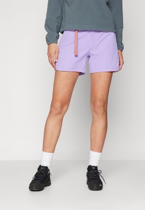 Person iført lavendelshorts, mørkegrå langærmet top, hvide strømper og sorte sneakers, der står mod en ensfarvet baggrund.