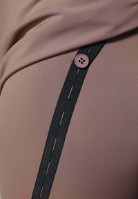 Un tissu brun clair à texture lisse présente une bande élastique noire et un bouton rond violet comme éléments de détail, créant une ligne de design épurée.