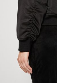 Manche d'une veste bomber en satin noir avec un design froncé et un poignet côtelé. Portée sur un vêtement en tissu noir texturé, mettant en valeur des textures lisses.