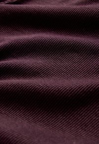Paarse corduroy stof met fijne verticale ribbels en zachte plooien die een getextureerd, golvend oppervlak creëren onder diffuus licht.