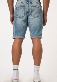Lichtblauwe denim shorts, met een opgerolde zoom, vijf zakken en een subtiele vervaagde textuur. Draagt witte hoge sneakers met sokken.