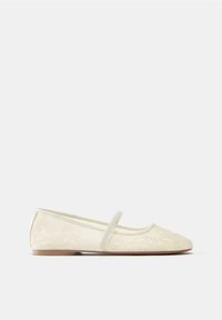Baletná obuv krémovej farby s kvetinovou výšivkou a jedným elastickým remienkom cez priehlavok, s plochou podrážkou, bočný pohľad na jednofarebnom pozadí.