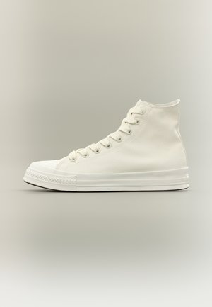 Kõrge säärega valge lõuendist toss valgete paelte ja kummist tallaga, nähtud külgprofiilist neutraalse tausta peal.