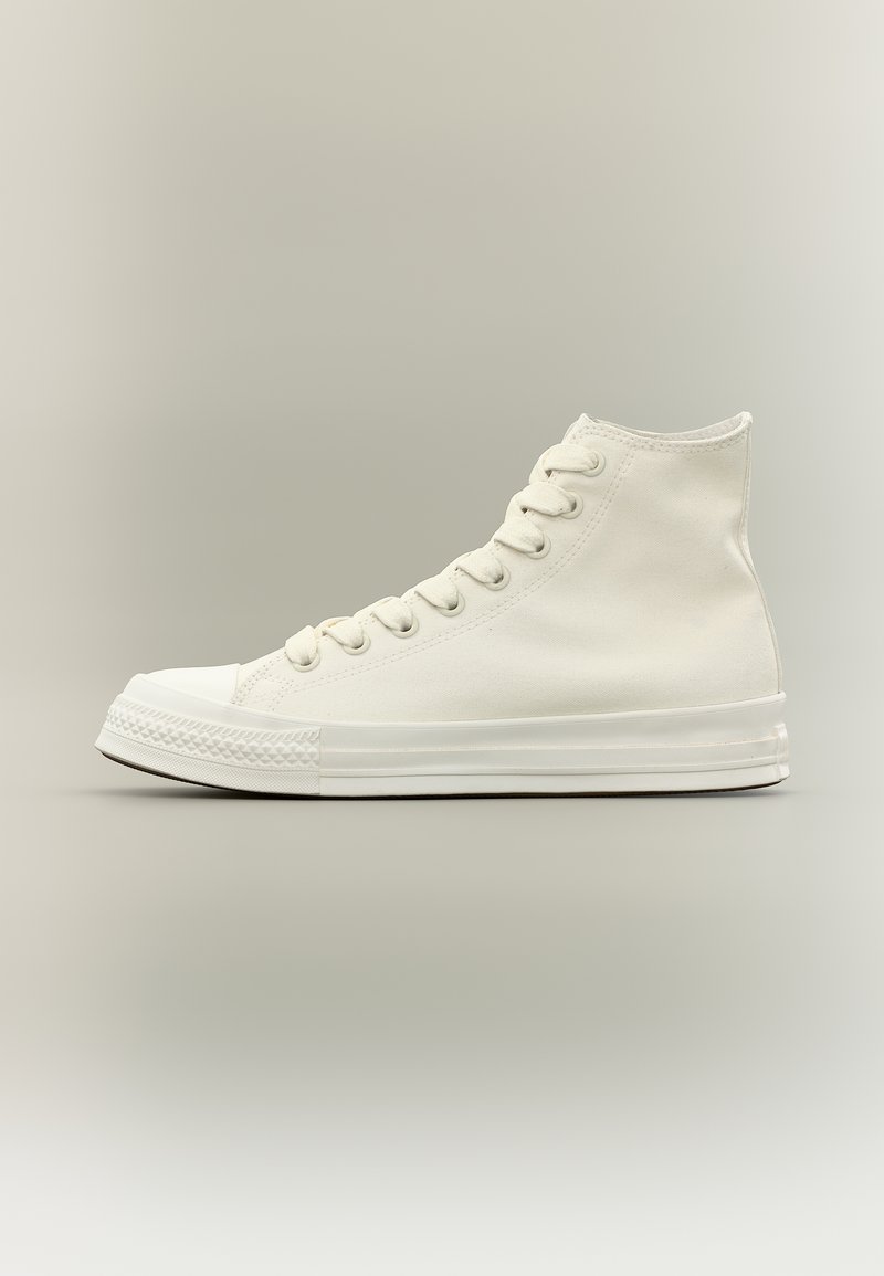 Aukštai einantis baltos drobės sportbačiai su baltais raišteliais ir guminėmis padais, parodyti šoniniu profiliu neutralioje fone.