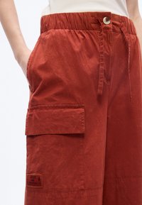 Pantalon cargo rouge rouille avec taille élastique, fermeture par bouton, grande poche à rabat sur la cuisse, et une main dans la poche latérale.