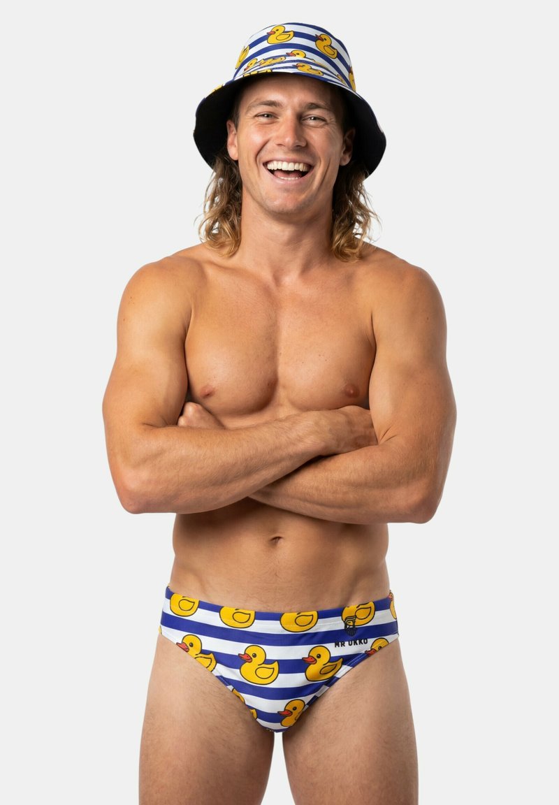 Uomo sorridente senza maglietta con capelli lunghi che indossa un cappello a secchiello e slip da bagno abbinati con motivo a paperelle, braccia incrociate su sfondo bianco.