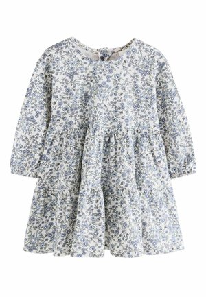 Giovane ragazza con vestito a fiori che tiene un grande fiore pallido con foglie marroni, guardando leggermente verso l'alto su uno sfondo chiaro e uniforme.