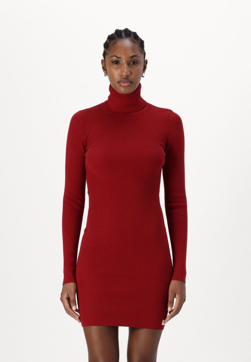 Sarkana ribota turtlenecka kleita ar garām piedurknēm, pieguļošs dizains un vidus augšstilba garums, ar mazu logo birku pie apakšmalas.