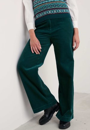 Pantalons en velours côtelé à jambes larges de couleur vert foncé avec une texture en relief, associés à un gilet en laine à motifs et des chaussures noires à semelles épaisses.