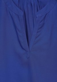 Modrá blůza s výstřihem do V, nařaseným výstřihem, hladkou texturou a krátkými rukávy. Uvnitř límce je ozdobné šití pro zvýraznění detailu.