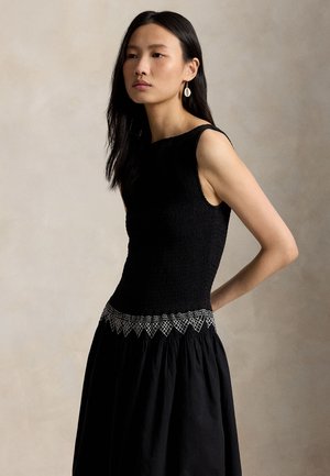 Femme aux longs cheveux noirs portant une robe noire sans manches avec un détail brodé blanc à la taille, se tenant devant un fond neutre.